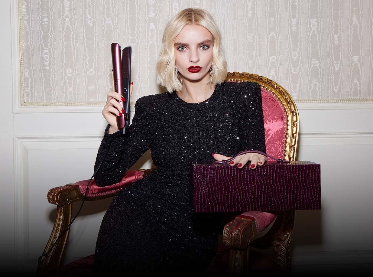 Model mit einem blonden welligen Bob hält das ghd gold Glätteisen in Kirschrot